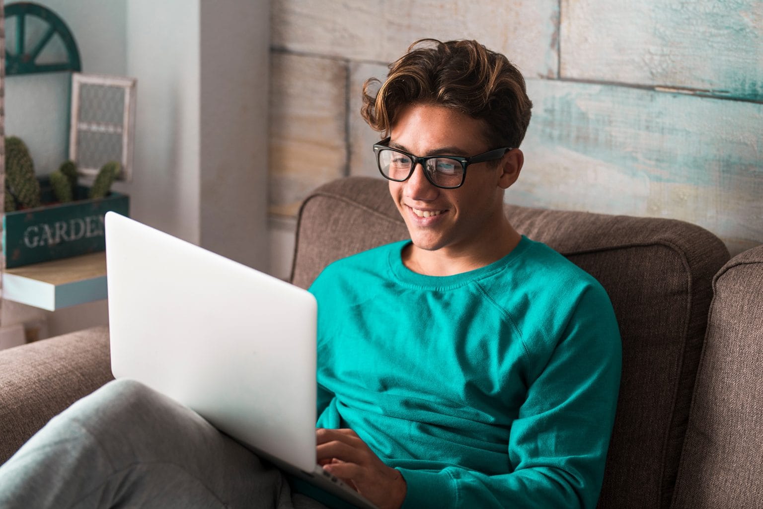 A man in glasses sitting in a chair in an apartment while smiling and looking at a laptop
